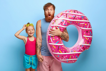Photo of stupefied bearded father holds hand of little daughter, carries inflated swimring, going together on beach. Glad redhaired girl wears goggles, enjoys summer rest with dad. Parenthood