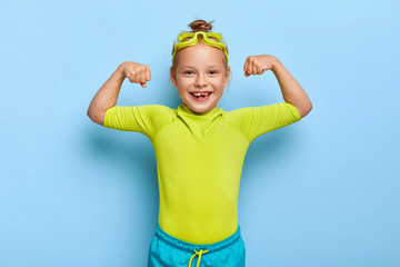 Positive little girl spends leisure time in water park, raises arms and shows muscles, ready for swim and dive, poses over blue background, wears goggles and swimsuit. Small kid has healthy lifestyle