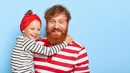Family and sincere emotions concept. Pleasant looking little female kid with red scraf around head, black and white striped jumper embraces father with big love has thruthful feelings enjoys free time