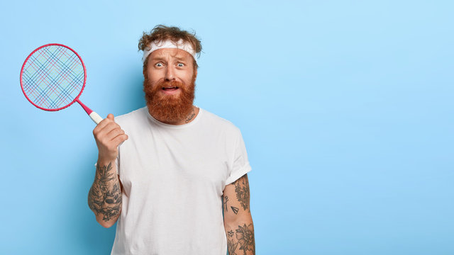 Horizontal shot of puzzled redhead man poses with tennis racket, being worn out of active game, wears casual clothes, poses over blue wall with empty space. Fatigue sportsman plays on tennis court