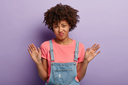 Thank yoy but no. Displeased dark skinned woman receivs disturbing suggestion, waves palms in rejection, grimaces unpleasantly, wears casual pink t shirt and dungarees, poses in studio alone