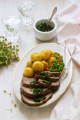 Slices of baked meat with potatoes and a garnish of peas and carrots on light background.