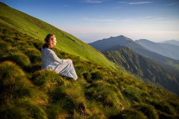 Young woman sitting on a rock and looking to the horizon. Happy traveler enjoying sunset view. Girl in long white dress in the mountains