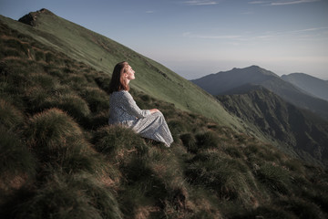 Young woman sitting on a rock and looking to the horizon. Happy traveler enjoying sunset view. Girl in long dress in the mountains