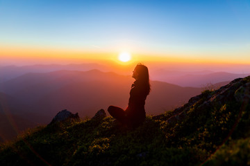 Girl on the background of mountain peaks. Woman hiking in mountains. Woman sitting on a rock over mountains under bright sky at sunset.