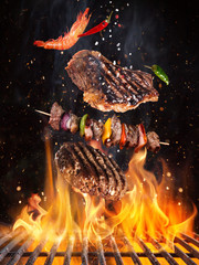 Tasty beef steaks and skewers flying above cast iron grate with fire flames. Freeze motion barbecue...
