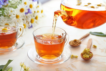 Pouring of tasty chamomile drink from teapot into cup on light table