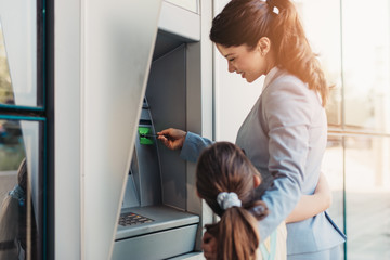 Young mother elegantly dressed with her daughter using ATM machine on city street.