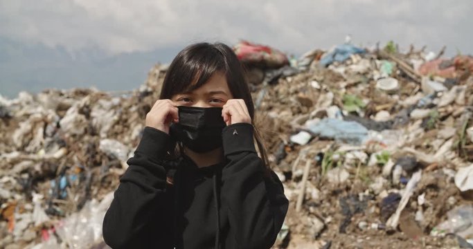 Little sad asian girl standing on disposal site, looking at camera with upset glare - ecological disaster concept portrait close up 4k