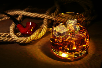 Glass with whiskey, ice cubes and smoking pipe on wooden background.