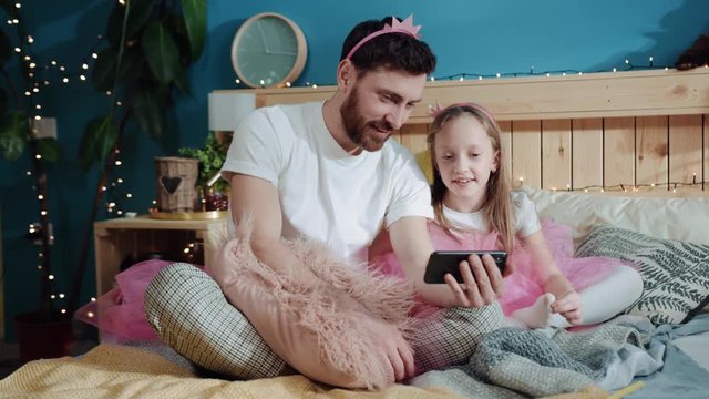 Family portrait of young daddy and his daughter dressed like fairies watching funny cartoons on smartphone sitting together in bedroom. Exciting family party. Joy and fun.