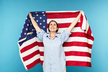 Happy young casual woman raising hand while holding large American flag on blue background