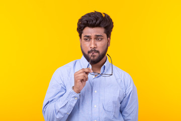 Young indian male professor with a beard in glasses and formal clothes is looking down thoughtfully posing on a yellow background. Concept of search and selection. Copy space