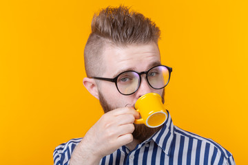 Cute confident young man drinking coffee from a little yellow mug while standing against a yellow background. Concept of morning coffee or espresso.