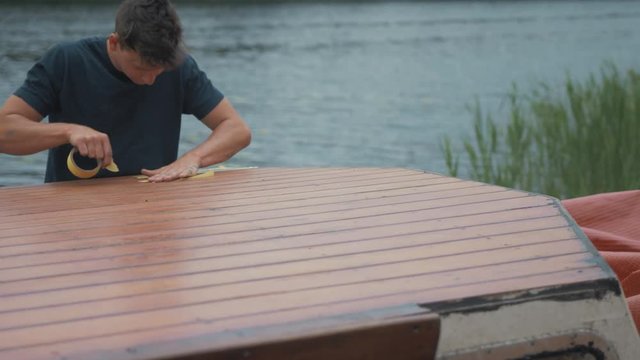 Young man masking wooden boat roof to seal with sika-flex