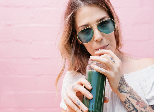 Young woman drinking a green juice with green sunglasses on a pink brick background