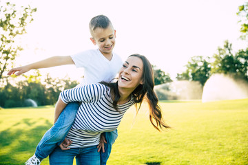 Happy beautiful young mother is playing with her little son while he is riding on her back and shouting for joy.