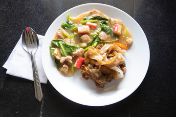Thai noodle with pork in gravy on white dish put on the black tile floor. with fork and spoon on white tissue.