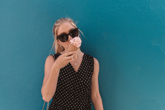 A beautiful young blonde woman eating a pink ice cream cone in the city