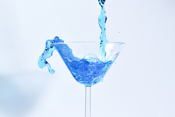 Cocktail with blue liquid in glass. Glass with blue water pouring with liquid with splashes and drops. Martini glass filling with alcohol with splashes on white background. Refreshing drink concept.