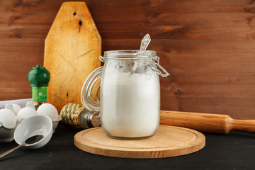 Open glass jar with white wheat flour and metal spoon in it on black wooden table among other kitchen stuff