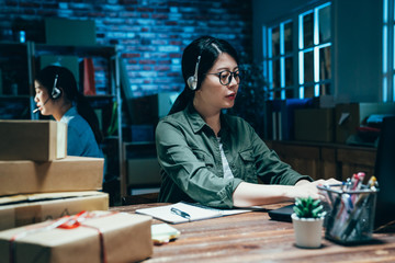 two female workers in warehouse wearing headset using laptop computer in dark night office. young girls partner business owner colleagues over worked in e commerce workplace customer support 24 hours