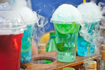 Multicolored fruit drinks close-up. Non-alcoholic drinks in plastic cups at the festival. Cocktails with juice and greens. White smoke is steaming with cocktails.