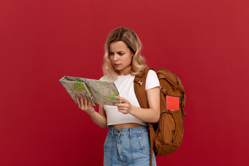 Thoughtful blond girl with curly hair in a white t-shirt looks at the map trying to find itinerary holding orange backpack