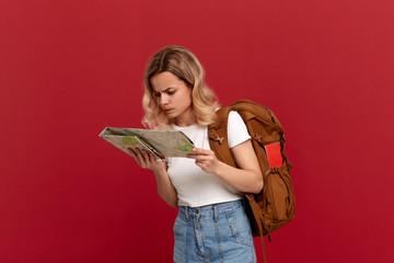 Thoughtful blond girl with curly hair in a white t-shirt looks at the map trying to find itinerary holding orange backpack
