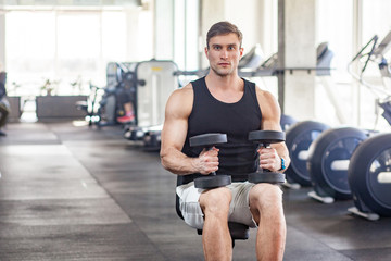 Obraz premium Portrait of young adult muscular built handsome athlete working out in a gym, sitting on a weightlifting machine and holding two dumbbell on the knees. indoor, looking at camera