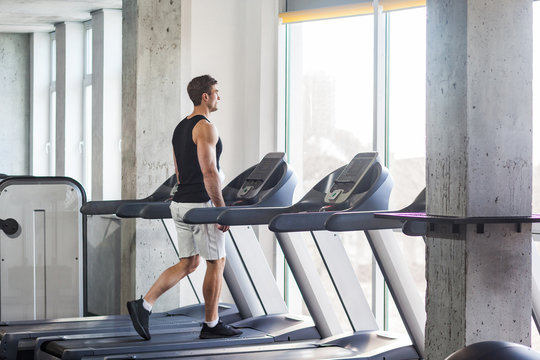 Side view of young adult sport man training at gym alone. Handsome male walking on treadmill and looking to window. Indoor, healthy fitness concept, athlete working out in a gym