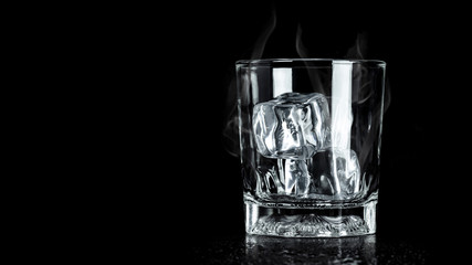 Empty glass with ice cube on dark background with cooling smoke and blank space. Ice blocks for beverage.