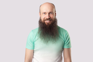 Portrait of happy satisfied middle aged bald man with long beard in light green t-shirt standing and looking at camera with toothy smile. indoor studio shot, isolated on grey background.