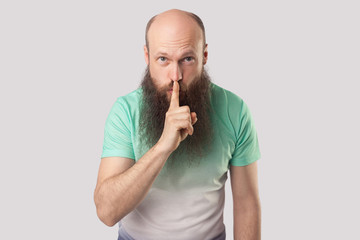 Be quiet. Portrait of serious middle aged bald man with long beard in light green t-shirt standing with finger on lips and showing silence sign. indoor studio shot, isolated on grey background.