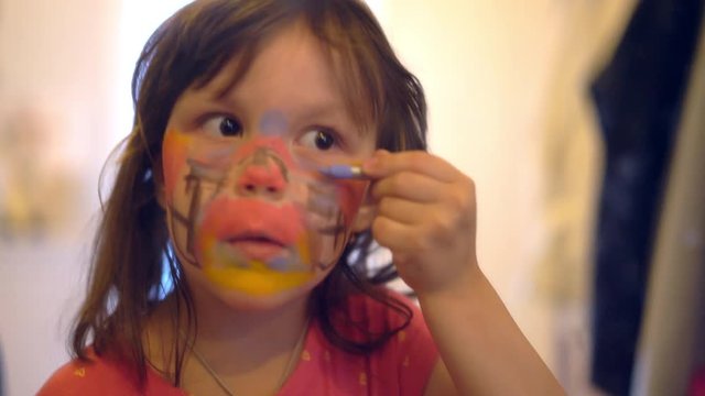 Portrait of little caucasian girl paints on face with face paint in front the mirror