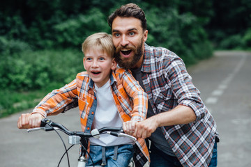 Handsome young dad and his cute little son with surprise laughing emotions are riding bikes in park, looking at camera and smiling.