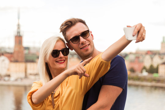 Portrait of the happy young couple who is standing in the embrace against the background of the city and river and doing selfie photo