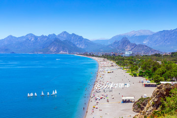 Panoramic view of town beach against the backdrop of mountains. Blue sea with sailboats and a picturesque beach. Holidays in the Mediterranean.