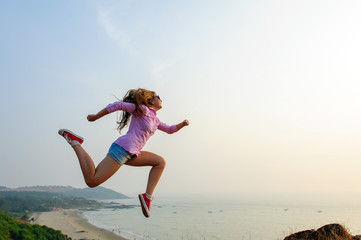 Beautiful happy young dark-haired woman jumps high and makes dynamic movements. Sporty girl in shirt, shorts and sneakers depicts running in the air on a hill against the sea. The concept of success.
