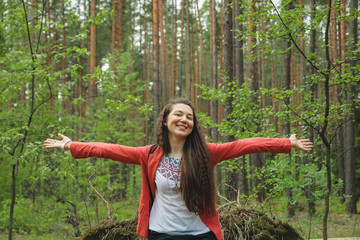 Young attractive smiling happy woman in red jacket with spread hands in spring forest among trees
