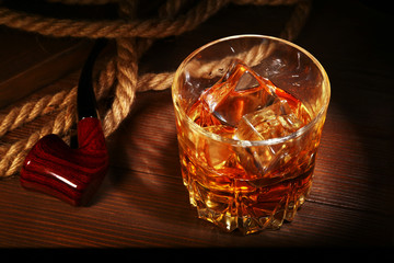 Glass with whiskey, ice cubes and smoking pipe on wooden background.