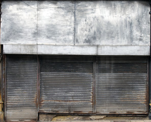 the front of an abandoned store on a street with closed dirty rusting metal shutters over the shop front and door