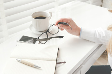 White office desk table with blank notebook, computer keyboard and other office supplies. Side view. Woman sitting at a desk, holding glasses in her hand