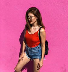 The model poses on the back of the pink wall.  Dressed in red top, blue shorts,  black backpack, denim sports shoes and   in fashionable mirror glasses. Photo taken on a beautiful sunny day