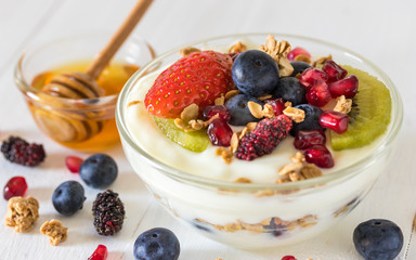 Plain yogurt with strawberry, blueberries, kiwi, granola, pomegranate in a glass bowl and honey on white wooden texture, healthy food and plant-based food concept