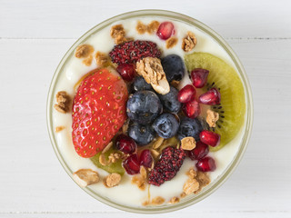 Top view of plain yogurt with strawberry, blueberries, kiwi, granola, pomegranate in a glass bowl and honey on white wooden texture,flay lay of healthy food and plant-based food concept