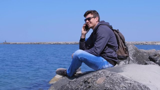 Man travaller calls smartphone sits on stone at sea background, side view. Young guy tourist talks mobile phone at sea beach. He is with backpack, wears jeans and sweatshirt in sunglasses