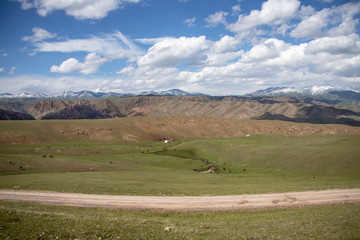 The road through the pasture between the hills with mountain ridges with snow covered peaks on the horizon. Travel. Kyrgyzstan
