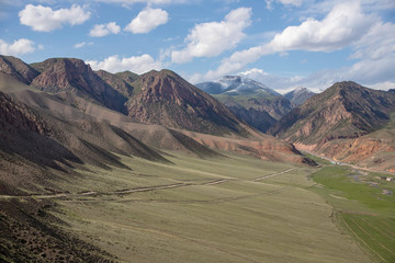 The road through the pasture between the hills with mountain ridges with snow covered peaks on the horizon. Travel. Kyrgyzstan