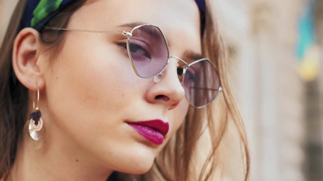 Outdoor close up portrait of young fashionable woman wearing trendy purple color sunglasses, colorful headband, earrings, posing outdoor, looking at camera